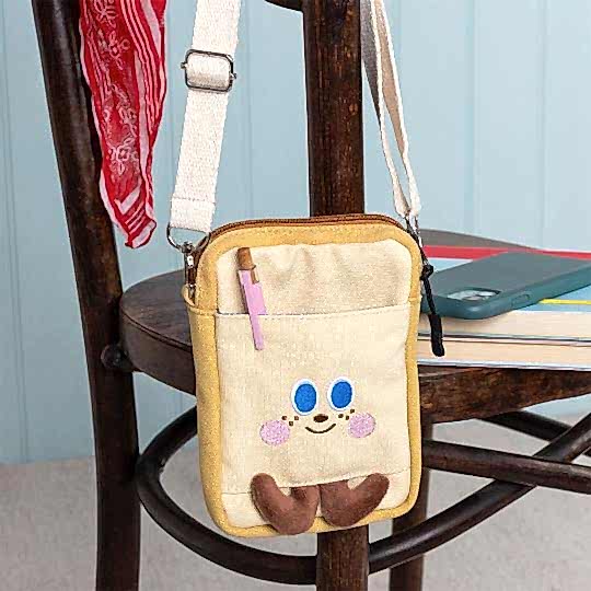 Beige and yellow crossbody bag with a front pocket featuring a smiling face design and two small brown plush feet at the bottom.
